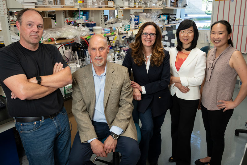 Five researchers pose together in a laboratory, standing and sitting among lab benches and scientific equipment